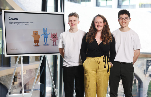 A photo of Daniel Valentine, Samantha Hodder and Alex Hung stood side by side next to a presentation slide. The slide is titled Chum and below has the text using education through stroytelling to help children during their cancer journey and shows three brightly coloured cartoon animals which represent the virtual chums