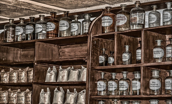 Clear glass jars with labels listing different chemicals are sat on top of old wooden shelves, reminiscent of an antique pharmacy