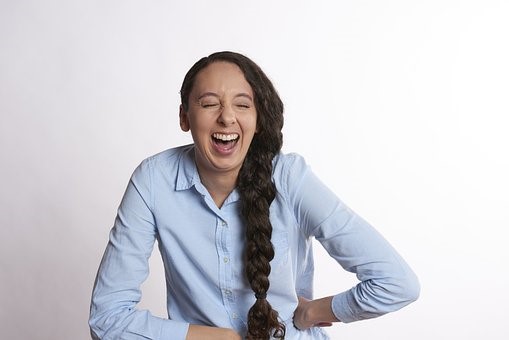 A woman facing the camera laughing with her eyes closed, with one hand on her hip and the other on her stomach