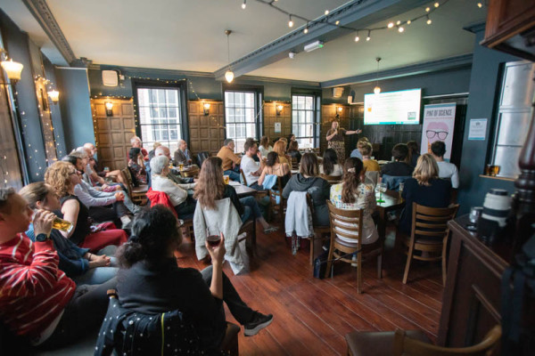 Audience members seated in a pub facing a speaker giving a presentation in front of a screen
